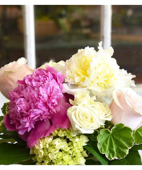 Peonies in shades of blush to strawberry pink are perfectly paired with roses, hydrangeas and seasonal foliage designed in a clear glass vase. 