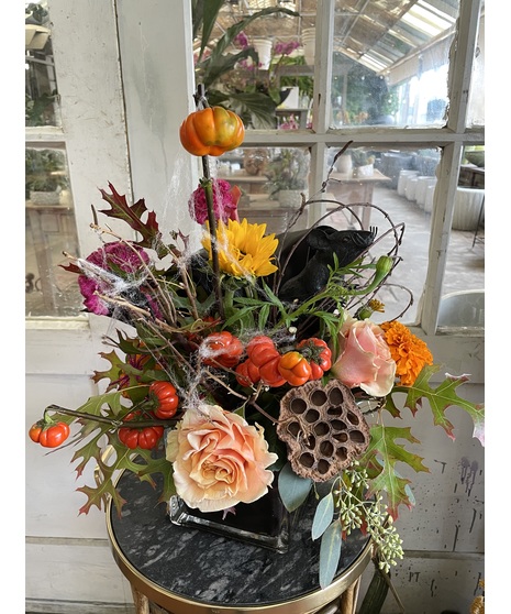 Halloween floral arrangement featuring orange mini pumpkins, peach roses, sunflowers, calla lilies, dried pods, branches, spiders, and a black bird in warm fall tones.