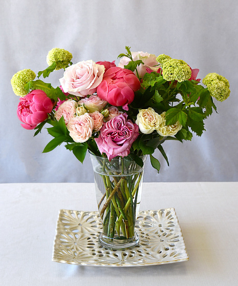 Peonies in shades of blush to strawberry pink are perfectly paired with roses, hydrangeas and seasonal foliage designed in a clear glass vase. 