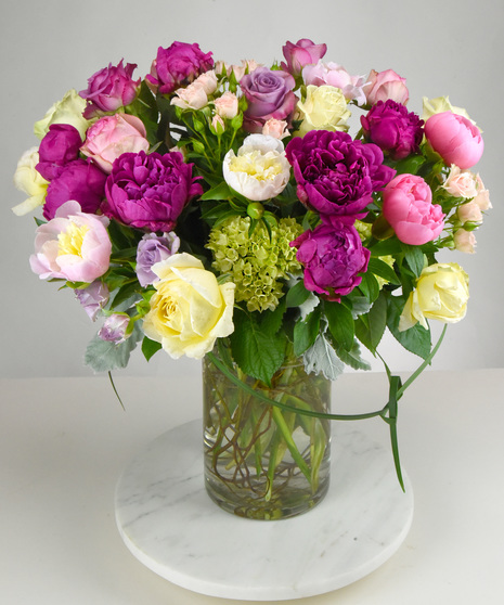 Peonies with a delicate scent is joined with fragrant roses in shades of pink and white  with seasonal foliage designed in a clear glass vase. 