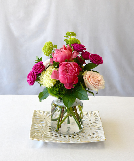 Peonies in shades of blush to strawberry pink are perfectly paired with roses, hydrangeas and seasonal foliage designed in a clear glass vase. 