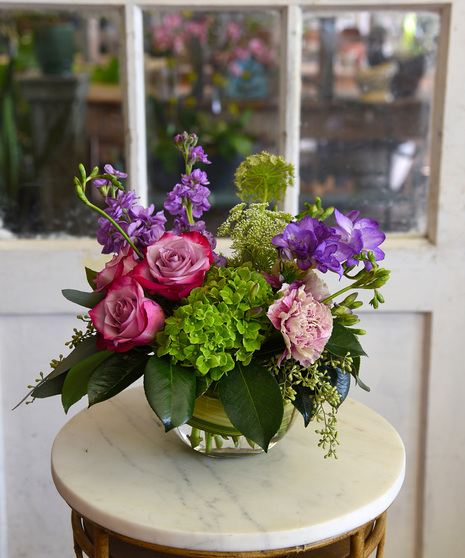 A contemporary design of lavender, pinks, cream and blue flowers like roses, hydrangea, carnations, Queen Anne's lace, with seasonal blooms and foliage n a  glass bubble bowl. 