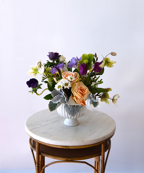 Garden-style floral arrangement with peach roses, purple tulips, lavender stock and freesia, purple anemones, white chrysanthemums, and hydrangea in a classic compote vase.