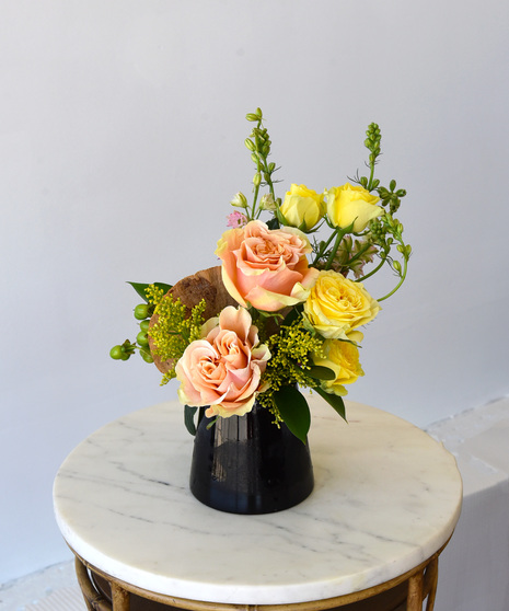 Contemporary floral arrangement with peach and yellow roses, aster, larkspur, and seasonal blooms in a black glass cone-shaped vase, with optional pink protea.
