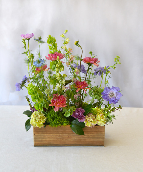 Joyful summer colors of orange, lavender, blues and pink in dahlias, larkspur, carnations, bells of Ireland and more in a natural rectangular wooden vessel.  