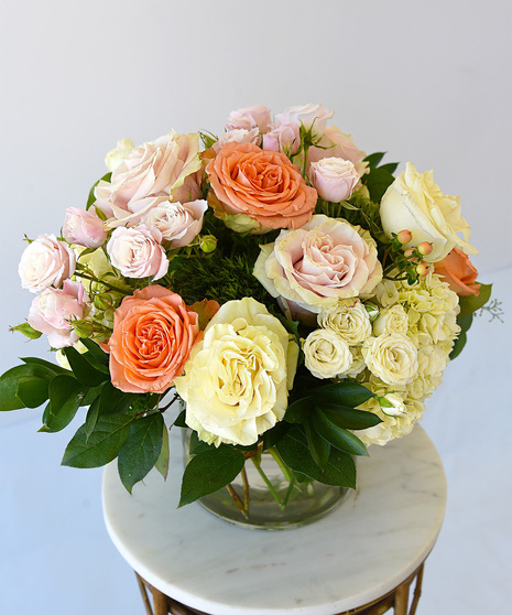 A lush collection of elegant colors of roses with spray roses in hues of peach, pink and white with hydrangea, and seasonal foliage in a glass vase.