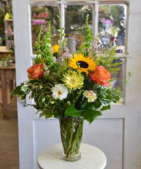 A colorful and vibrant summertime blooms with sunflowers, gerandos, snapdragons, roses, and gerberas among seasonal foliage handcrafted in a glass vase.  