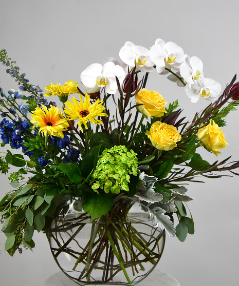 A contemporary design of phalaenopsis orchids, roses, delphinium hydrangea, gerbera intertwined with seasonal blooms and foliage in cool blues and white and sunny yellows.