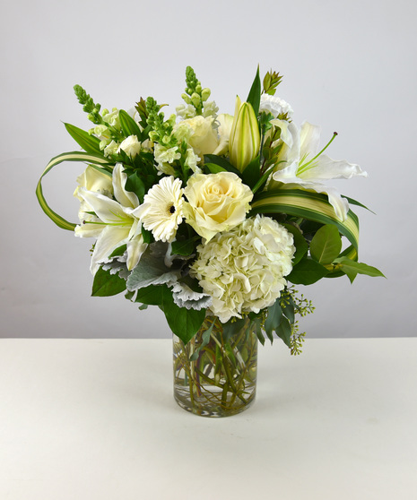 White lilies, roses and snapdragons among hydrangea, carnations and seasonal foliage in vase.   The grand is with phalaenopsis orchids, lush roses and additional flowers. 