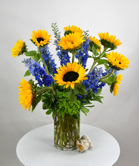 Bright yellow sunflowers and blue delphinium with foliage designed in a tall glass cylinder vase