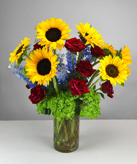 Bright yellow sunflowers and blue delphinium with foliage designed in a tall glass cylinder vase