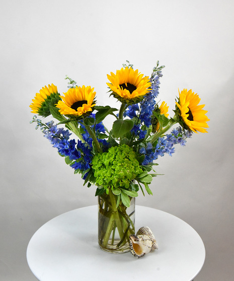 Bright yellow sunflowers and blue delphinium with foliage designed in a tall glass cylinder vase