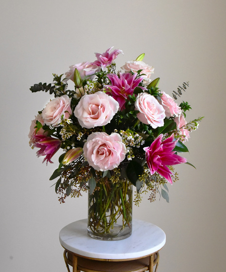 Pastel pink roses with ruffled multi-petal vibrant pink lilies with seasonal foliage white hydrangea,  snapdragons and wax flower presented in a glass vase. 