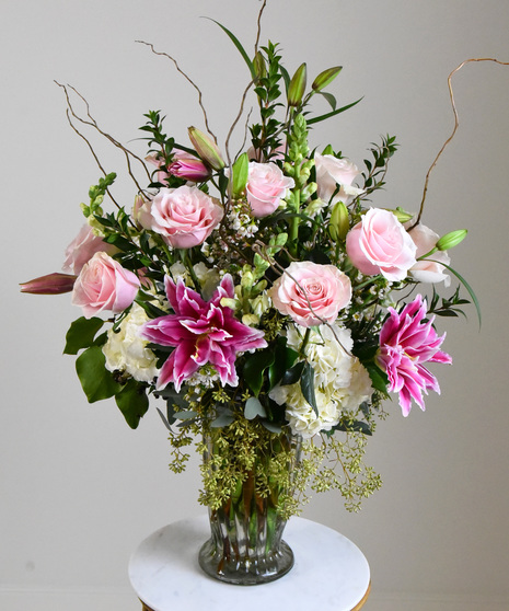 Pastel pink roses with ruffled multi-petal vibrant pink lilies with seasonal foliage white hydrangea,  snapdragons and wax flower presented in a glass vase. 