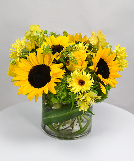 Sunflowers with yarrow, bupleurum and sometimes gerbera in a glass vase.  Also offered in a ceramic vase in a classic and stylized design of sunflowers and yarrow.