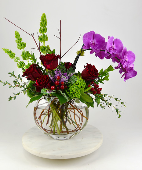  A contemporary design of phalaenopsis orchids, roses, hydrangea intertwined with seasonal blooms and foliage designed in a a clear glass sphere vase.