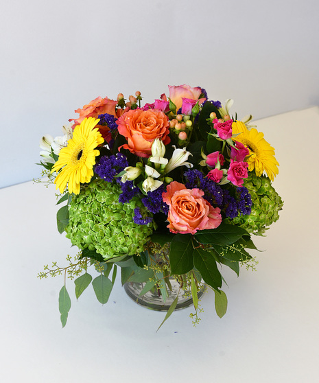 Cheerful design of roses, daisy, hydrangea in shades of yellow, orange, purple in glass vase.