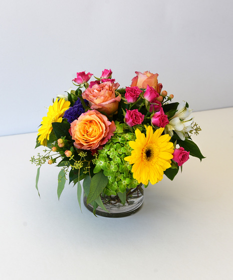 Cheerful design of roses, daisy, hydrangea in shades of yellow, orange, purple in glass vase.