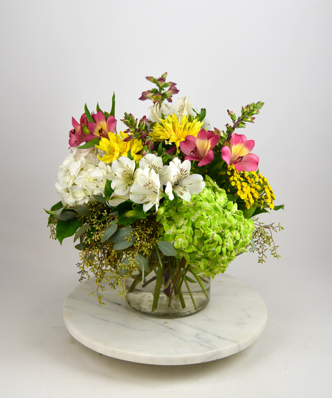 Cheerful design of roses, daisies, hydrangeas in shades of yellow, orange, pinks and blues in glass vase.