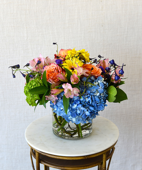 Cheerful design of roses, daisies, hydrangeas in shades of yellow, orange, purple in glass vase.