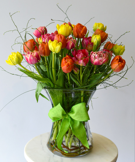 Multi-petal tulips in yellow and orange and bright pink or purple. They bloom among seasonal foliage in a glass vase with a spring color ribbon.  