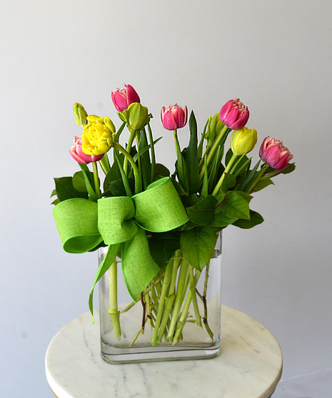 Multi-petal tulips in yellow and orange and bright pink or purple. They bloom among seasonal foliage in a glass vase with a spring color ribbon.  