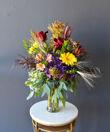 Modern artistry of Autumn colors of red roses, yellows sunflowers, shades of purple hydrangea and blush pink alstroemeria are surrounded by the foliage of fall.