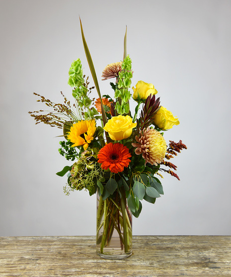 Splash your surroundings with modern artistry of festive Autumn colors of burgundy, pumpkin orange, warm yellows surrounded by the foliage of fall designed in a glass vase.  