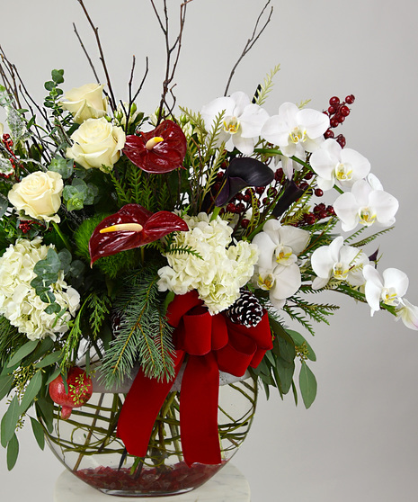 Blooms of creamy roses, hydrangea and deep purple calla lilies,  red berries, pinecones, seasonal foliage and a holiday red velvet bow. Designed in a glass sphere vase.