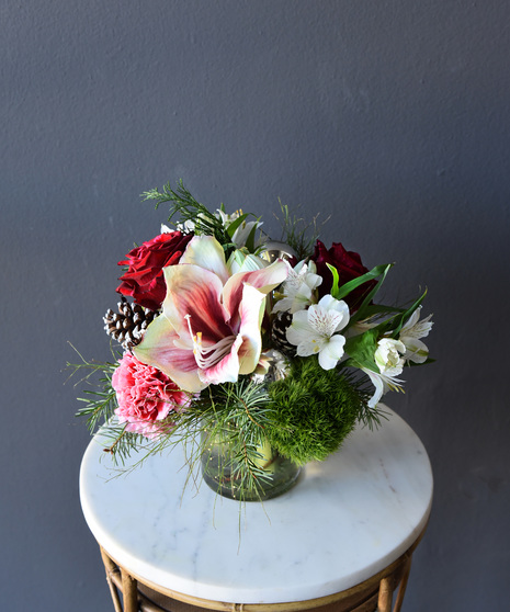 A palette of festive blooms amarylis, red roses, variegated carnations, hydrangea, hypericum berries, lisianthus, pinecones, seasonal foliage and designed in a glass vase.
