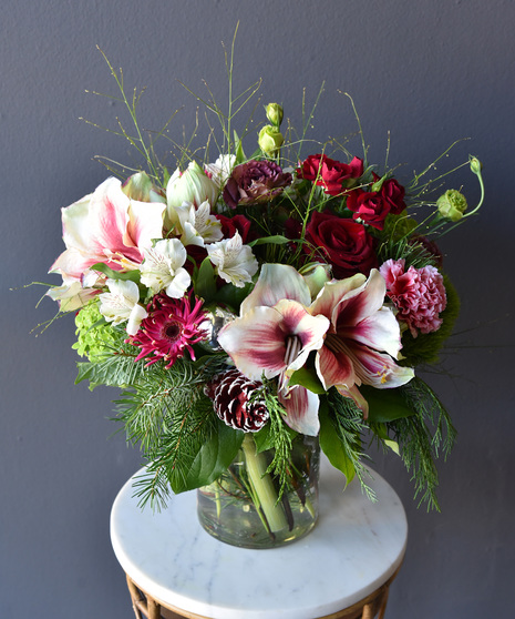 A palette of festive blooms amarylis, red roses, variegated carnations, hydrangea, hypericum berries, lisianthus, pinecones, seasonal foliage and designed in a glass vase.