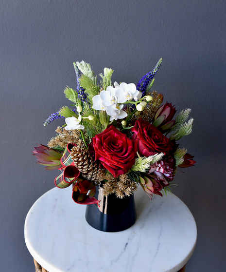 Deep-hued roses and orchids mingle with lavender blue veronica, winter greens, pinecones, and a graceful holiday bow, all set in a sleek black glass vase.